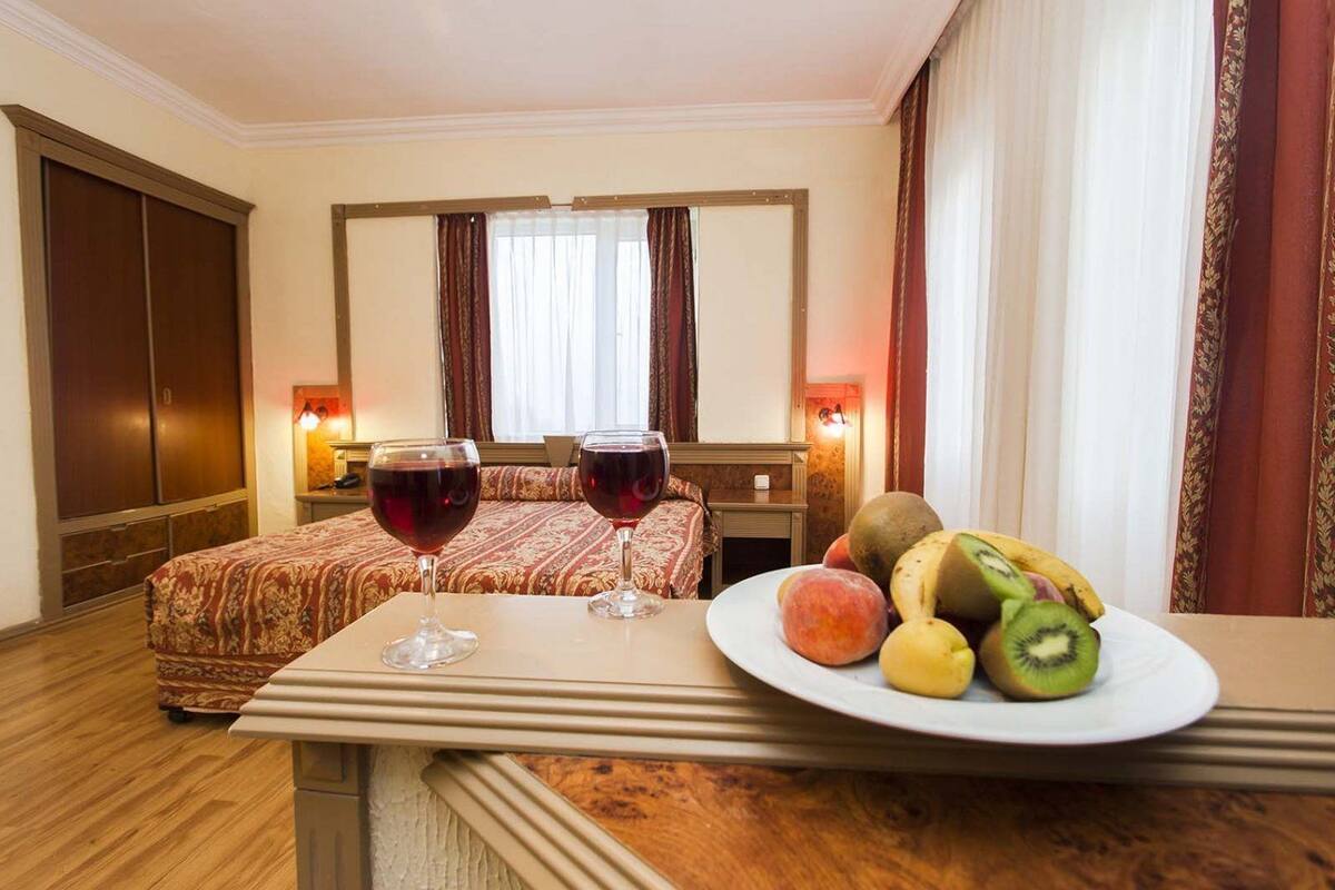 A hotel room with a bed, two bedside tables, a wooden nightstand, a plate of fruit, and two glasses of red wine.