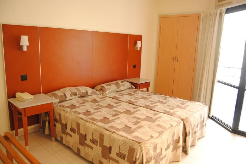 Habitació d'hotel amb dos llits, un armari de fusta, una taula petita amb una làmpada i una finestra amb cortines.