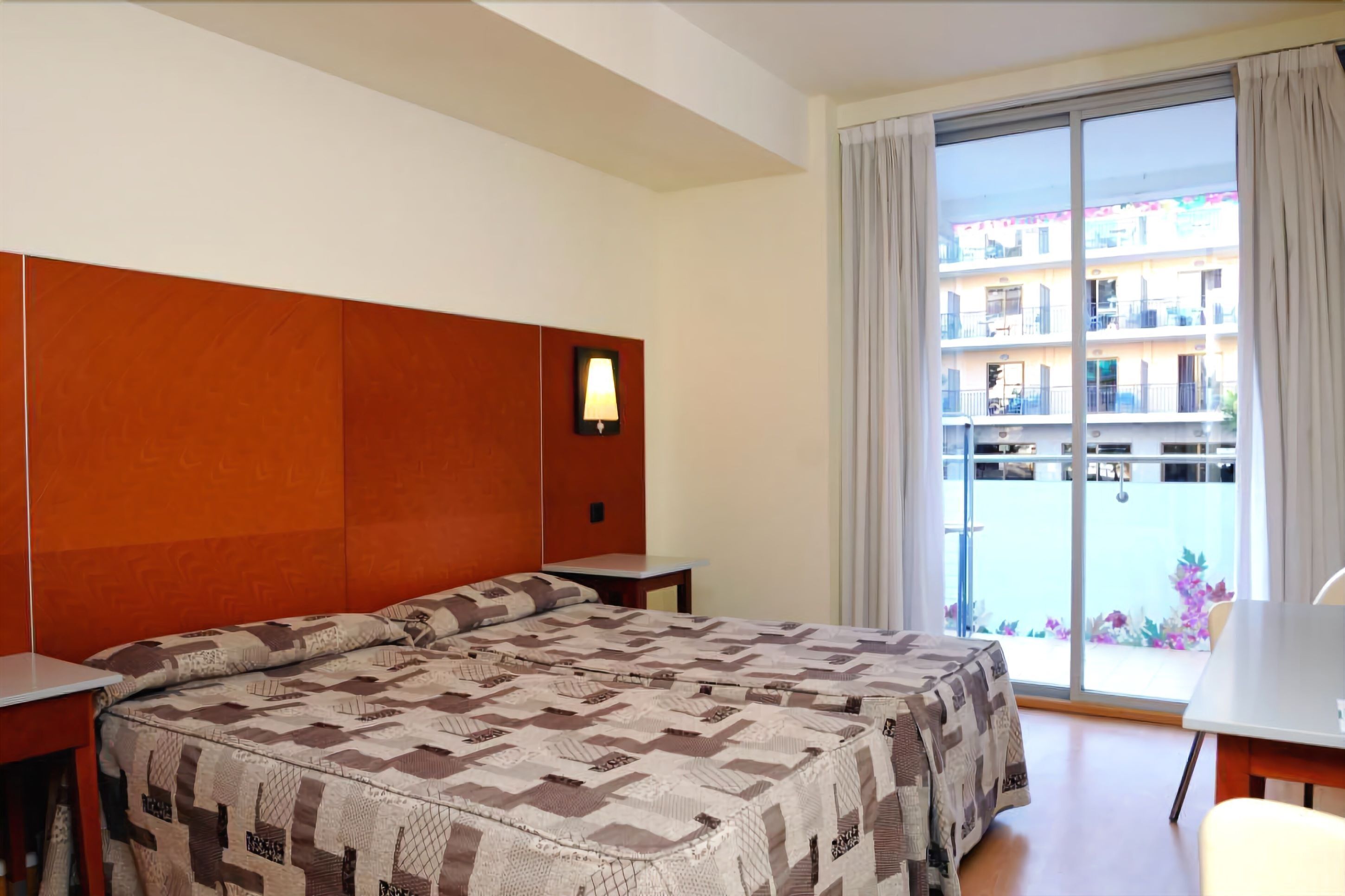 A hotel room with a large bed, a nightstand, a desk, and a window with a view of a building and balcony.