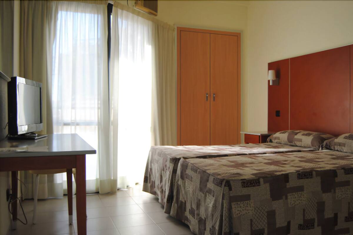 Habitación de hotel con cama, televisor en un escritorio, armario y ventana con cortinas.