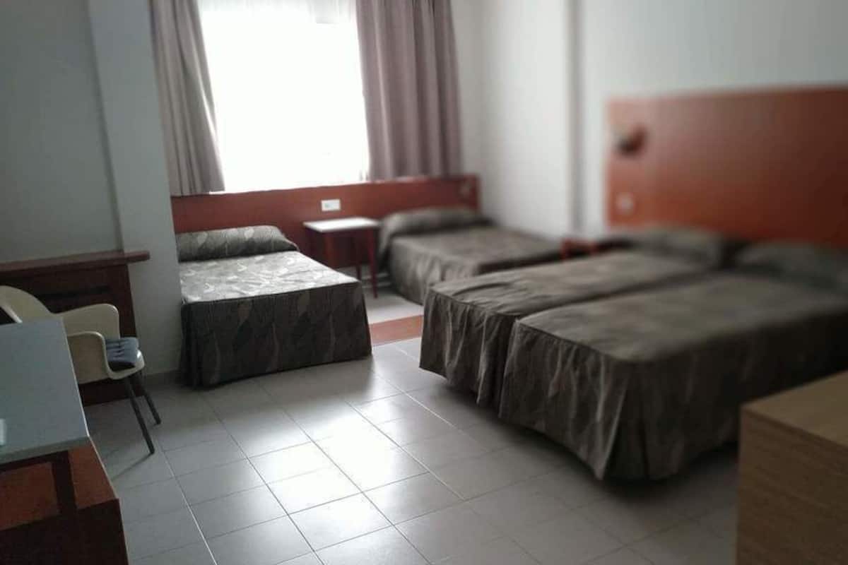 Habitación de hotel con dos camas individuales, una mesita, una silla y un ventanal con cortinas.