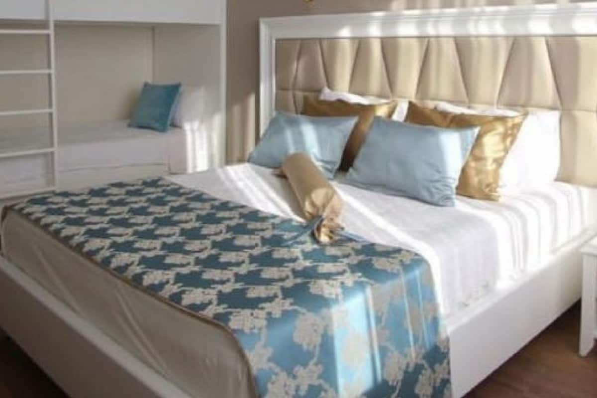 A hotel room with a bunk bed, a large bed with a patterned bedspread, and a bedside table with a lamp.