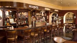 Un área de bar con sillas y mesas de madera, un mostrador con una caja registradora y estantes con botellas.
