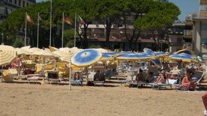 Een strand met rijen ligbedden en parasols, een houten looppad en gebouwen op de achtergrond.