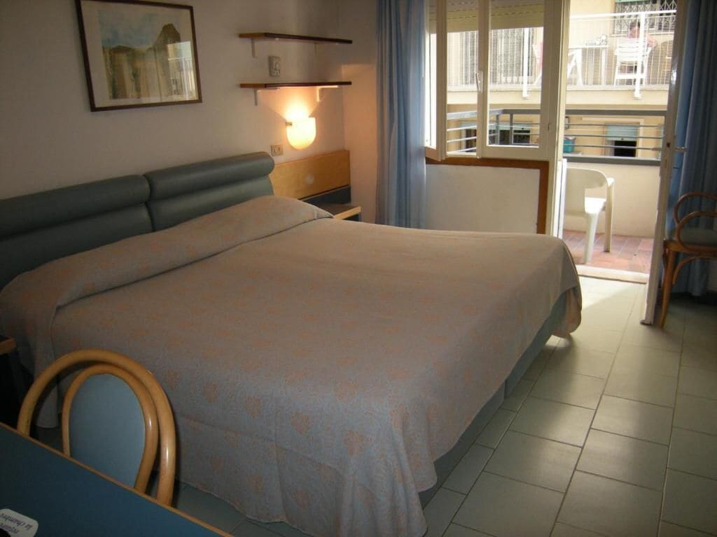 Un dormitorio con cama, mesita de noche, una silla, ventana con cortinas y balcón con barandilla.