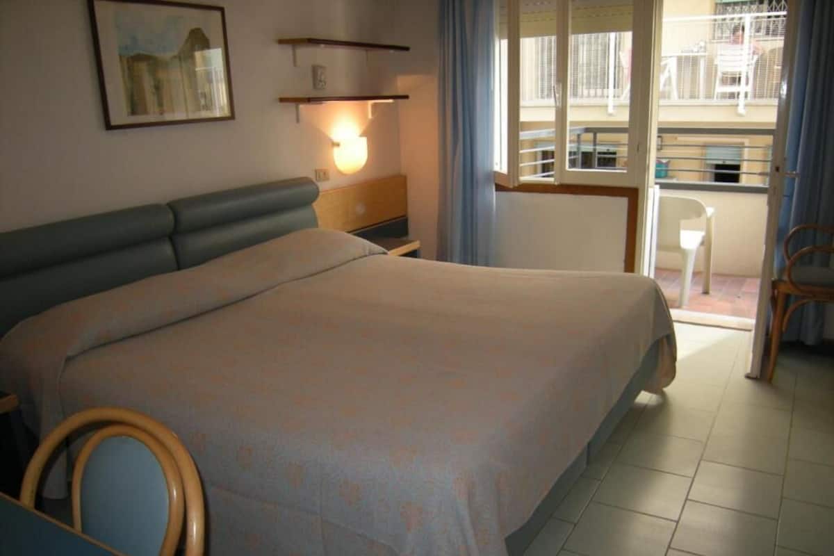 A bedroom with a bed, a bedside table, a chair, a window with curtains, and a balcony with a railing.