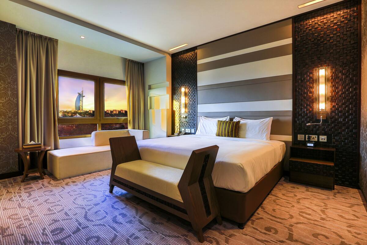 A modern hotel room with a large bed, a sofa, a view of a cityscape, and a decorative wall with lights.