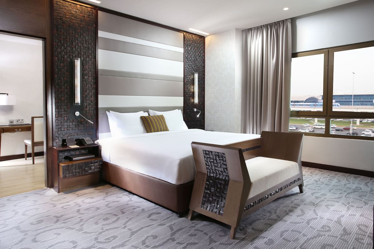 A modern hotel room with a large bed, a desk, a chair, and a view of the airport.