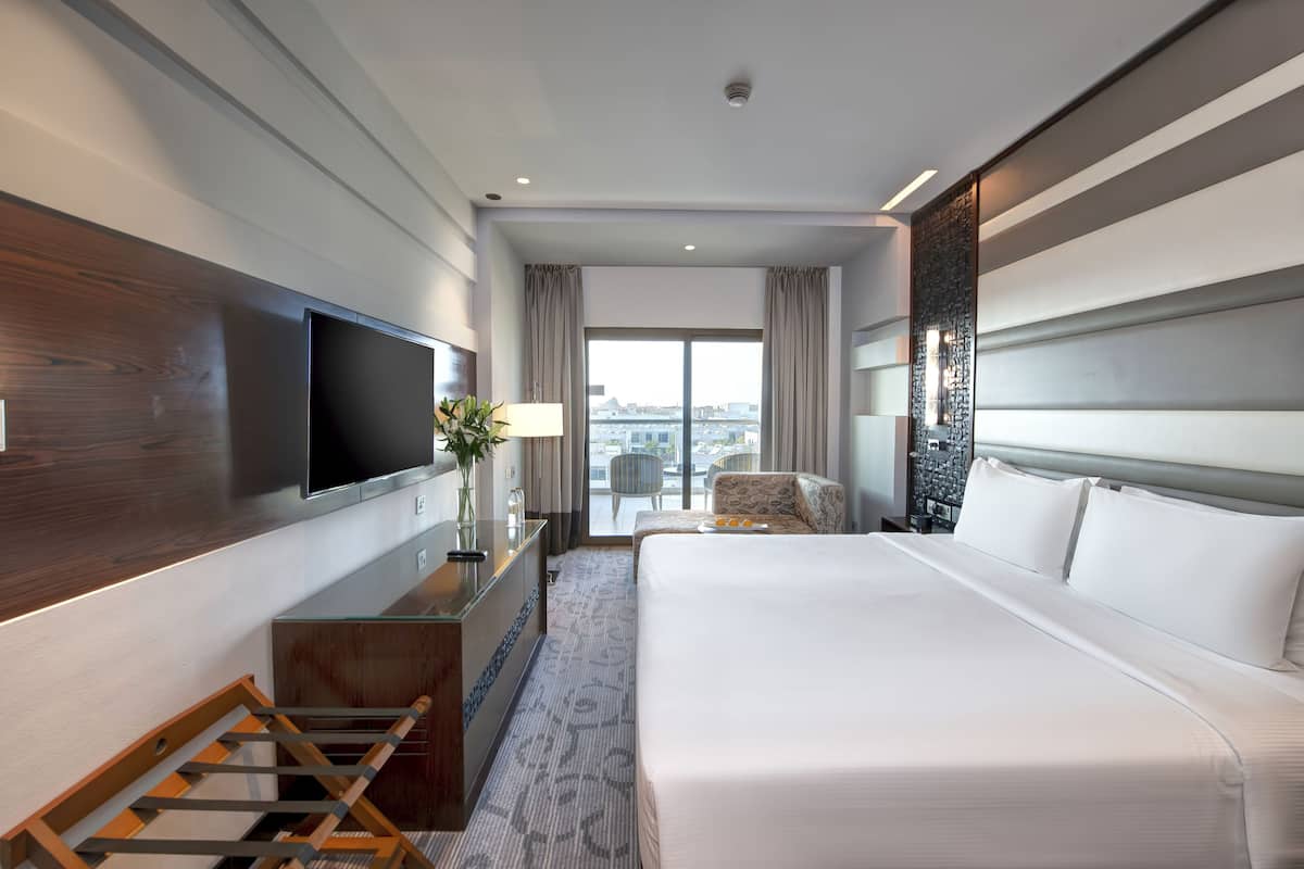 A modern hotel room with a large bed, a flat-screen TV mounted on the wall, a desk with a lamp, and a view of the city through the window.