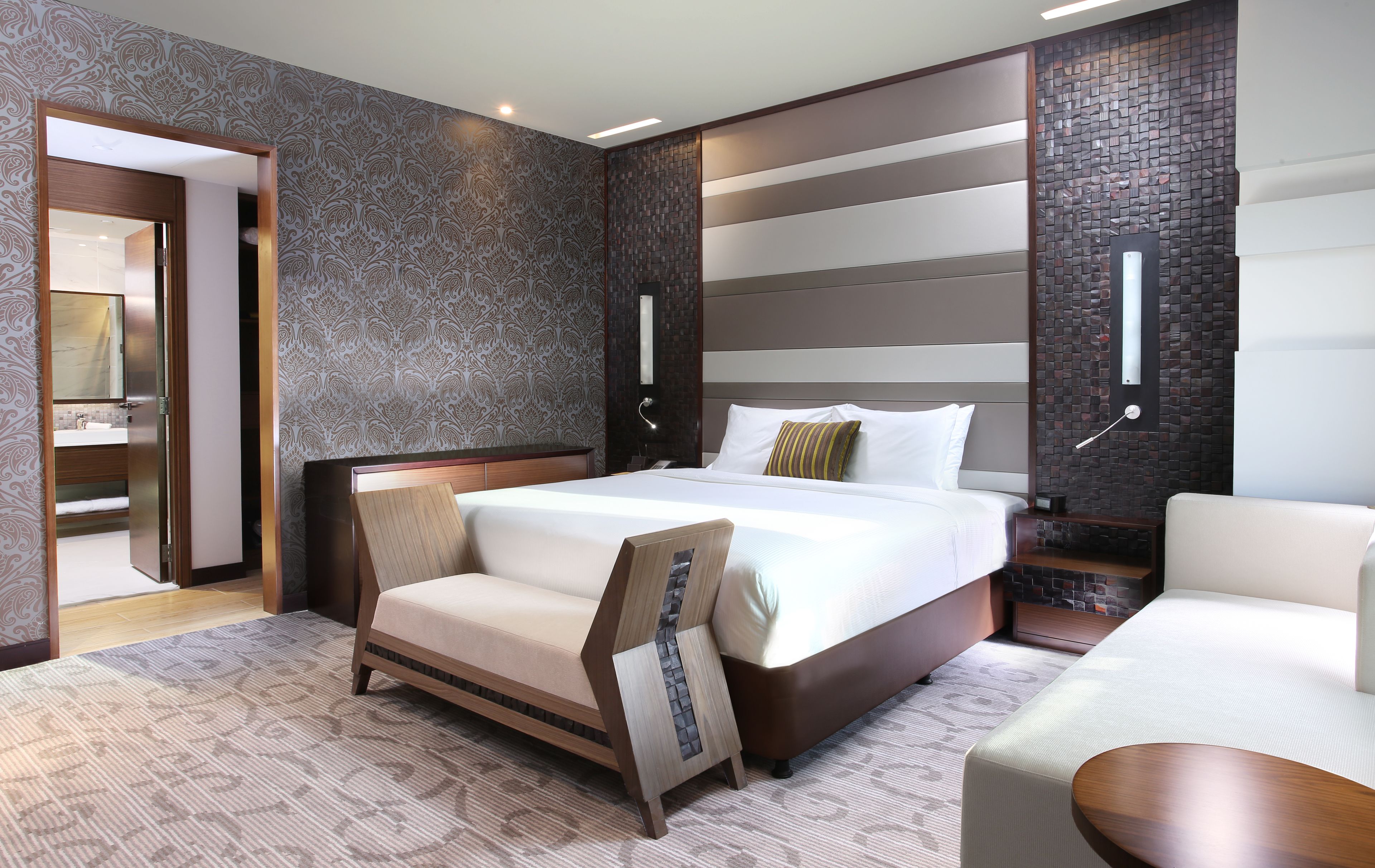 A modern hotel room with a large bed, a sofa, a small table, and a bathroom visible through an open door.