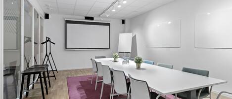 Una sala de conferencias con una mesa blanca, sillas blancas, una pantalla de proyector y un ventanal grande.