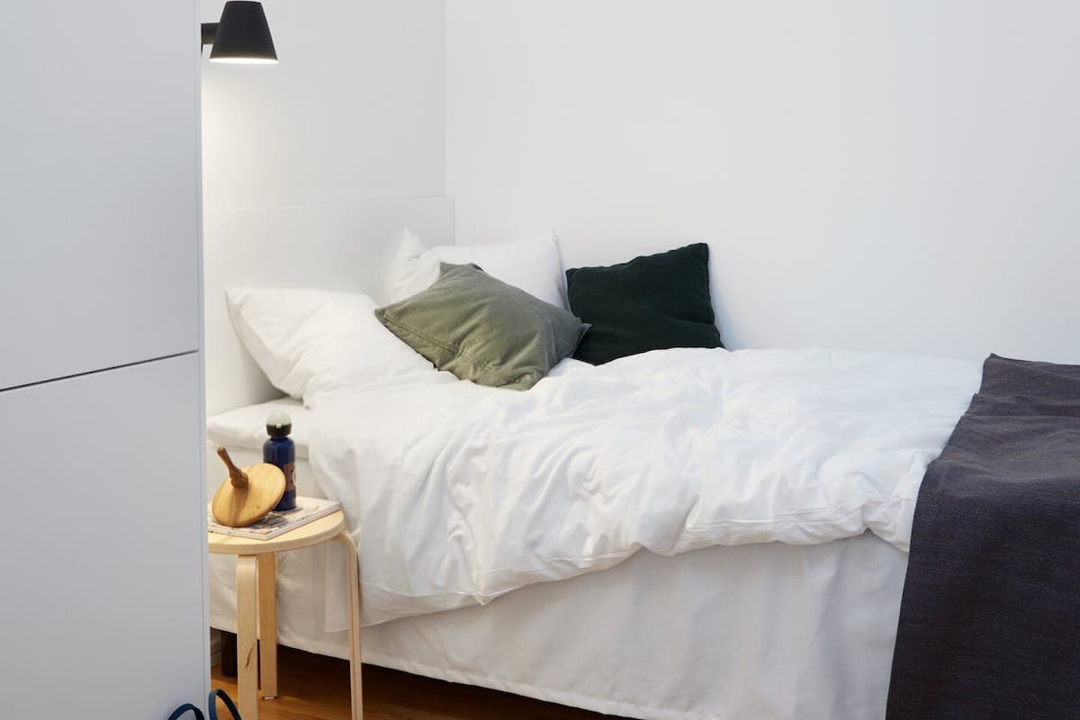Una cama pequeña y ordenada con ropa de cama blanca y un edredón oscuro, una lámpara de noche, una mesita de noche de madera con una guitarra y una botella, y una mochila azul en el suelo.