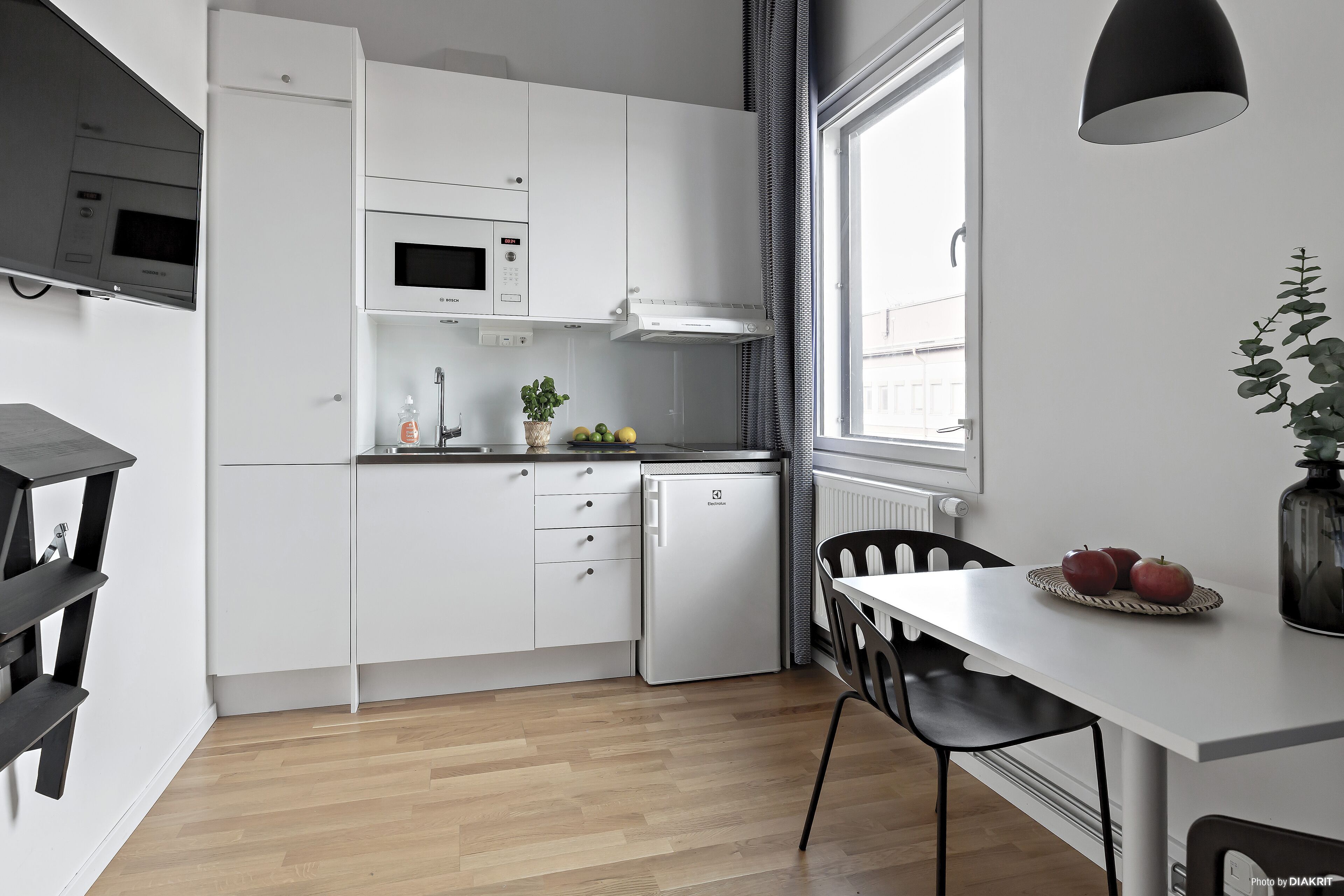 A compact kitchen with white cabinetry, a built-in microwave, and a small refrigerator. There is a dining table with chairs, a potted plant, and a fruit bowl on the counter.