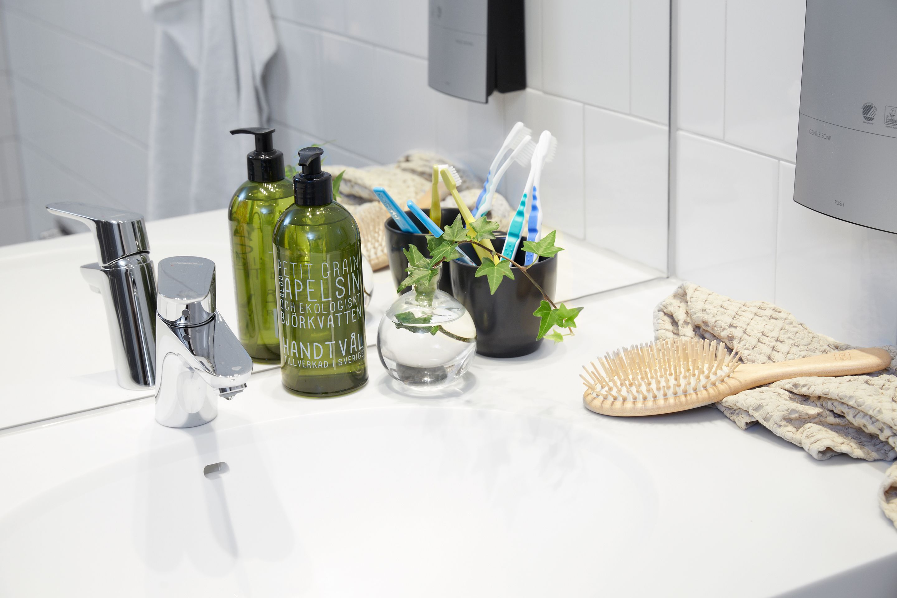 Un lavamanos con grifo cromado, una botella verde, un vaso transparente y un portalapices para cepillos de dientes con cepillos.