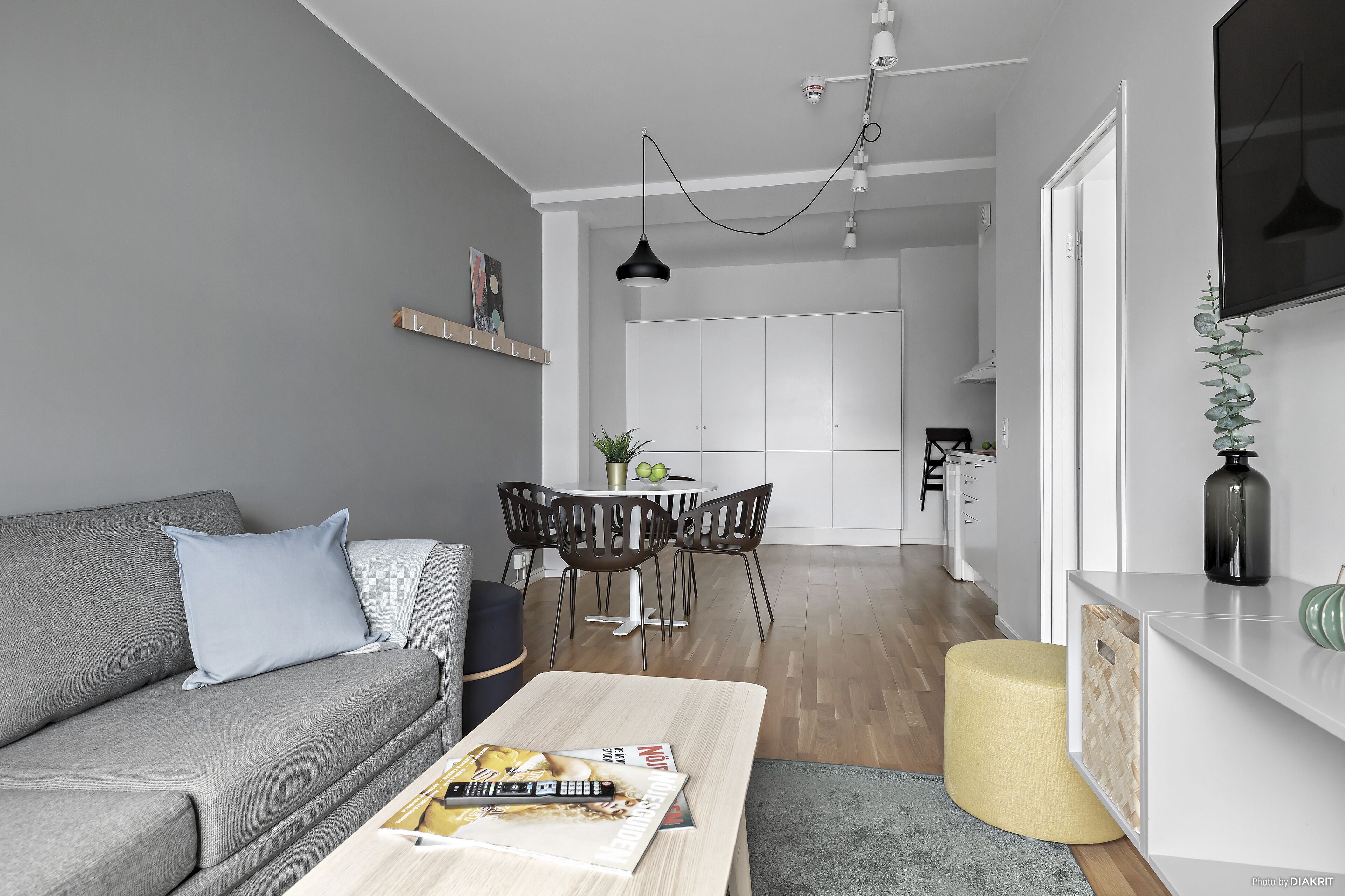 A modern living room with a grey sofa, a coffee table with magazines, a dining area with a white table and chairs, and a kitchen in the background.