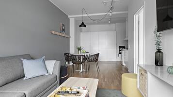 A modern living room with a grey sofa, a coffee table with magazines, a dining area with a white table and chairs, and a kitchen in the background.