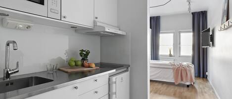 Una cocina moderna equipada con microondas, estufa y lavaplatos. Hay un pequeño comedor con una mesa y sillas. La habitación tiene una cama con una manta rosa y un televisor montado en la pared.