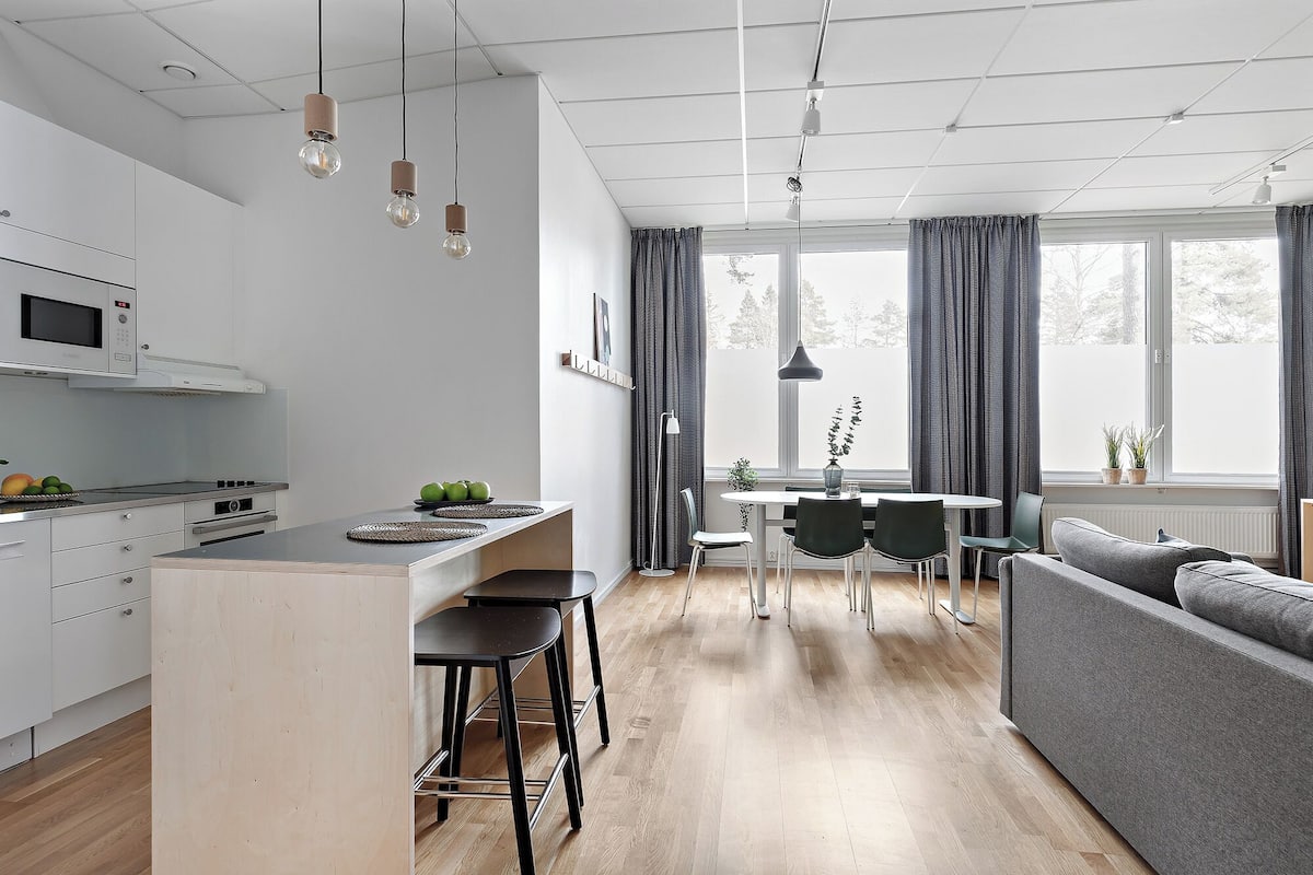 Una cocina moderna con una isla central, un comedor con una mesa redonda y sillas, y un sofá gris en una habitación luminosa con ventanas amplias.