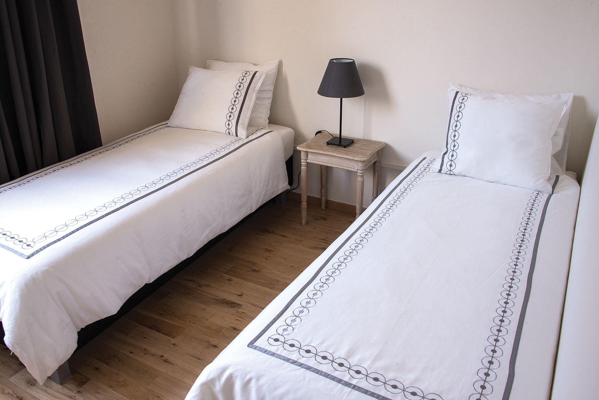 Ein Hotelzimmer mit zwei Einzelbetten, einem kleinen Nachttisch mit Lampe und einem Fenster mit Verdunkelungsvorhängen.
