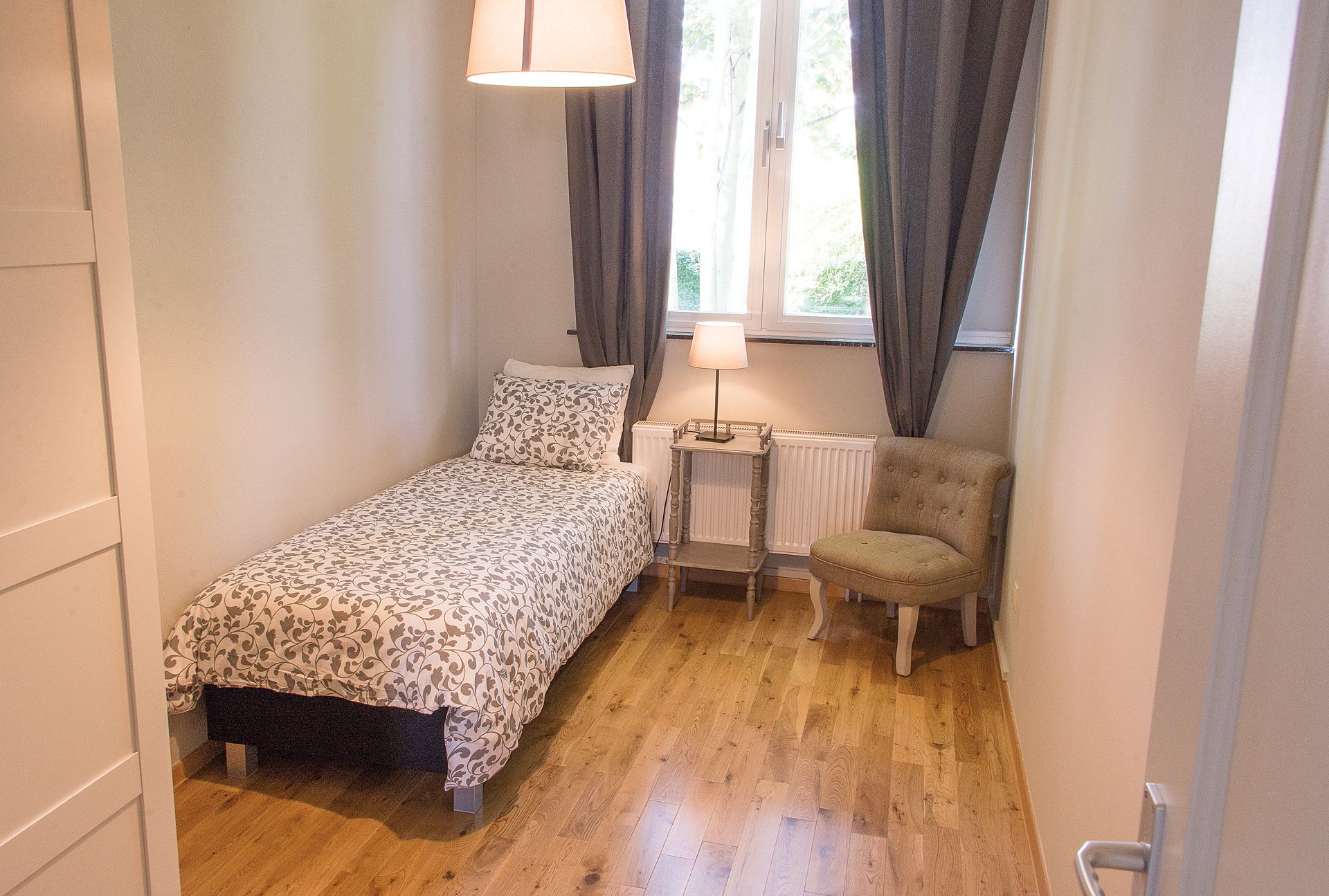 Een kleine, knusse slaapkamer met een eenpersoonsbed, een stoel, een tafel met een lamp en een raam met gordijnen.