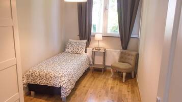 Een kleine, knusse slaapkamer met een eenpersoonsbed, een stoel, een tafel met een lamp en een raam met gordijnen.