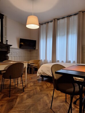 Een hotelkamer met een houten visgraatvloer, een bed, een bureau met stoel, een aan de muur bevestigde televisie en een groot raam met gordijnen.