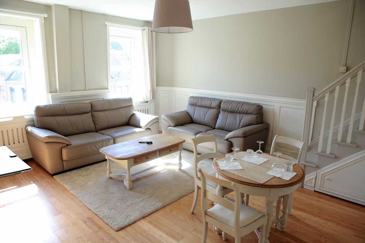 A living room with two beige sofas, a wooden coffee table, a round dining table with chairs, and a staircase in the background.