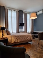 Habitación de hotel con cama, escritorio con lámpara, televisor montado en la pared, sofá de cuero y un ventanal con cortinas.