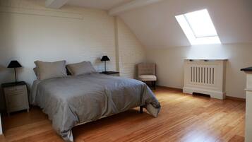 Een slaapkamer met een schuine kap, dakraam, bed met grijze dekbedovertrek, nachtkastje met lamp, stoel en radiator.