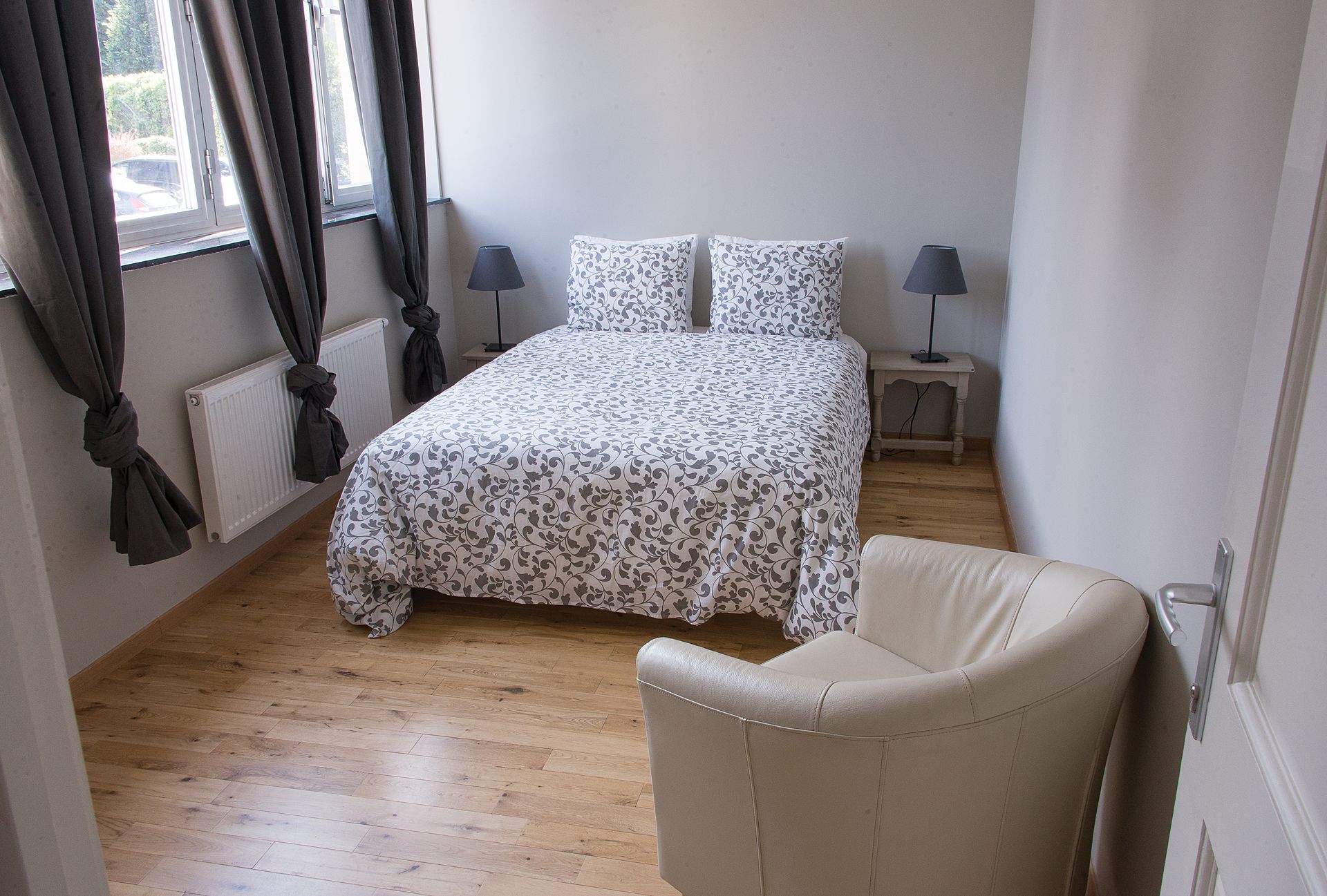 Un dormitorio con cama, dos lámparas de noche, una silla y una ventana con cortinas.