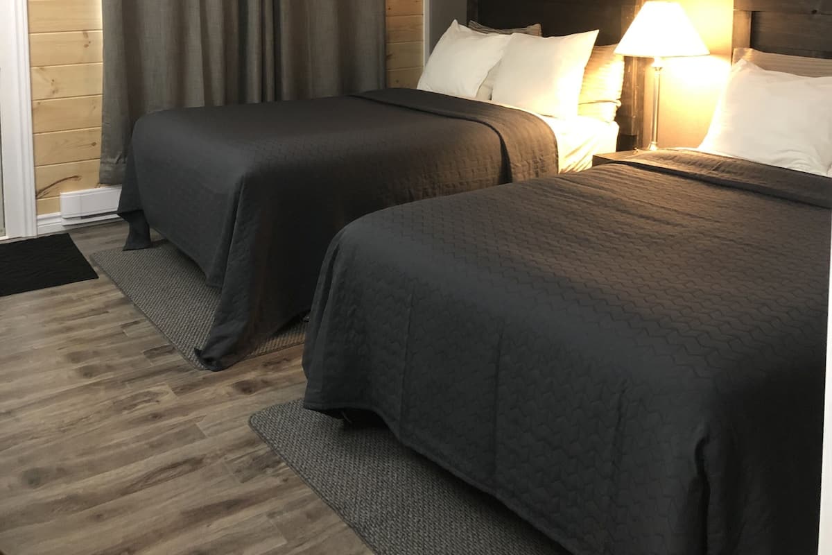 A hotel room with two beds, a wooden wall, a grey curtain, and a nightstand with a lamp.