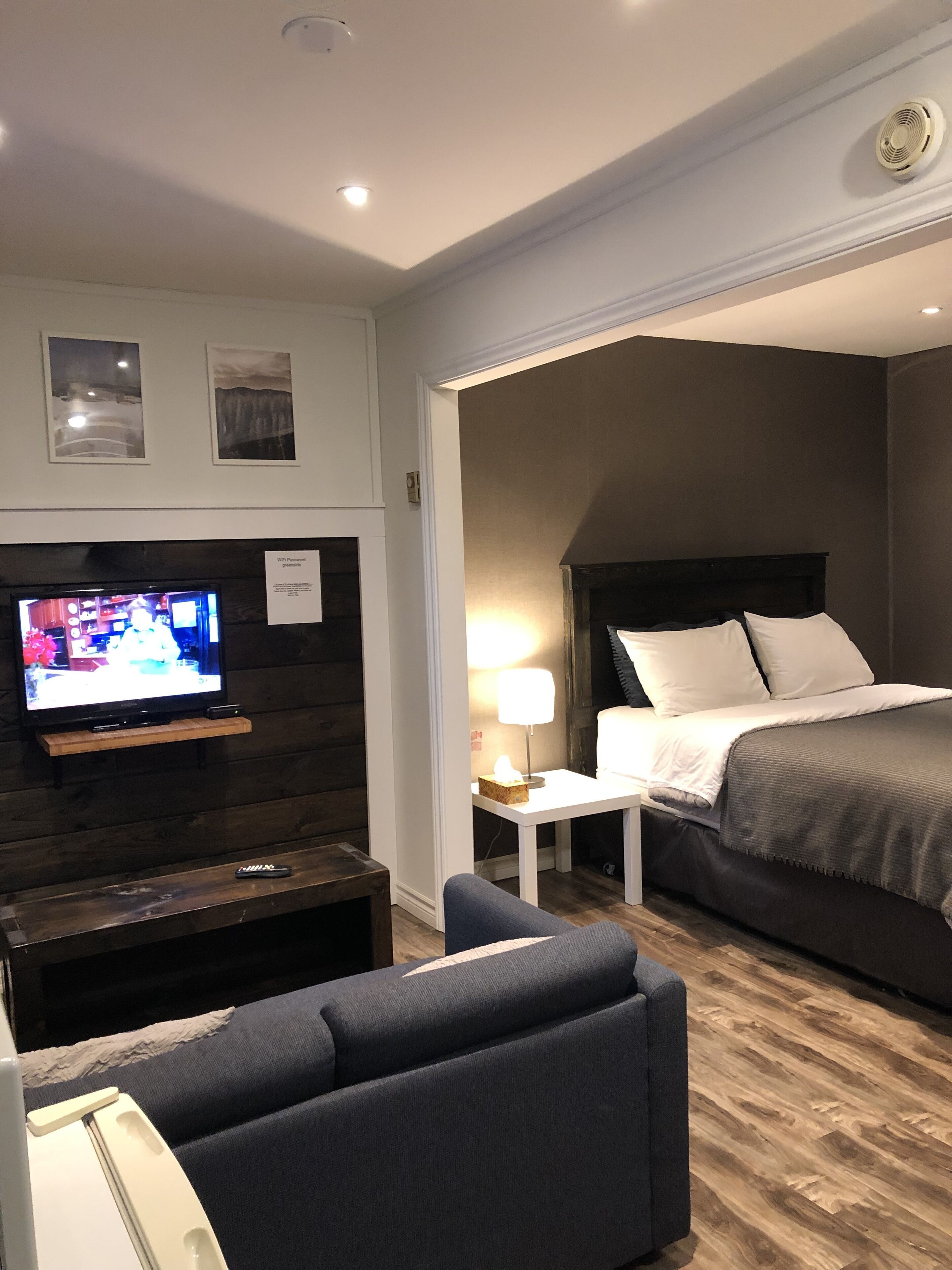 Una habitación de hotel con un televisor en un estante de madera, una cama con ropa blanca, una mesita de noche con una lámpara y un sillón de color oscuro.