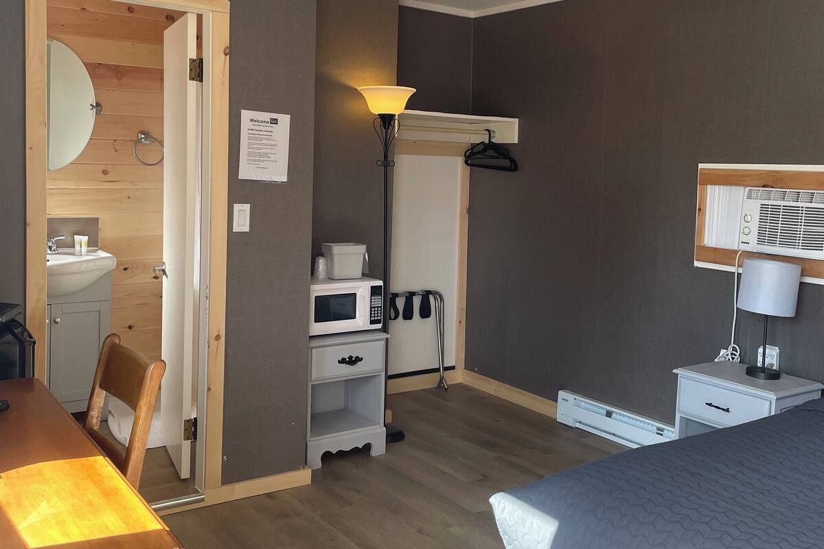 A hotel room with a wooden floor, a bed with a patterned bedspread, a desk with a chair, a bedside table with a lamp, a microwave, a small cabinet, a hanging coat rack, and a door leading to a bathroom.