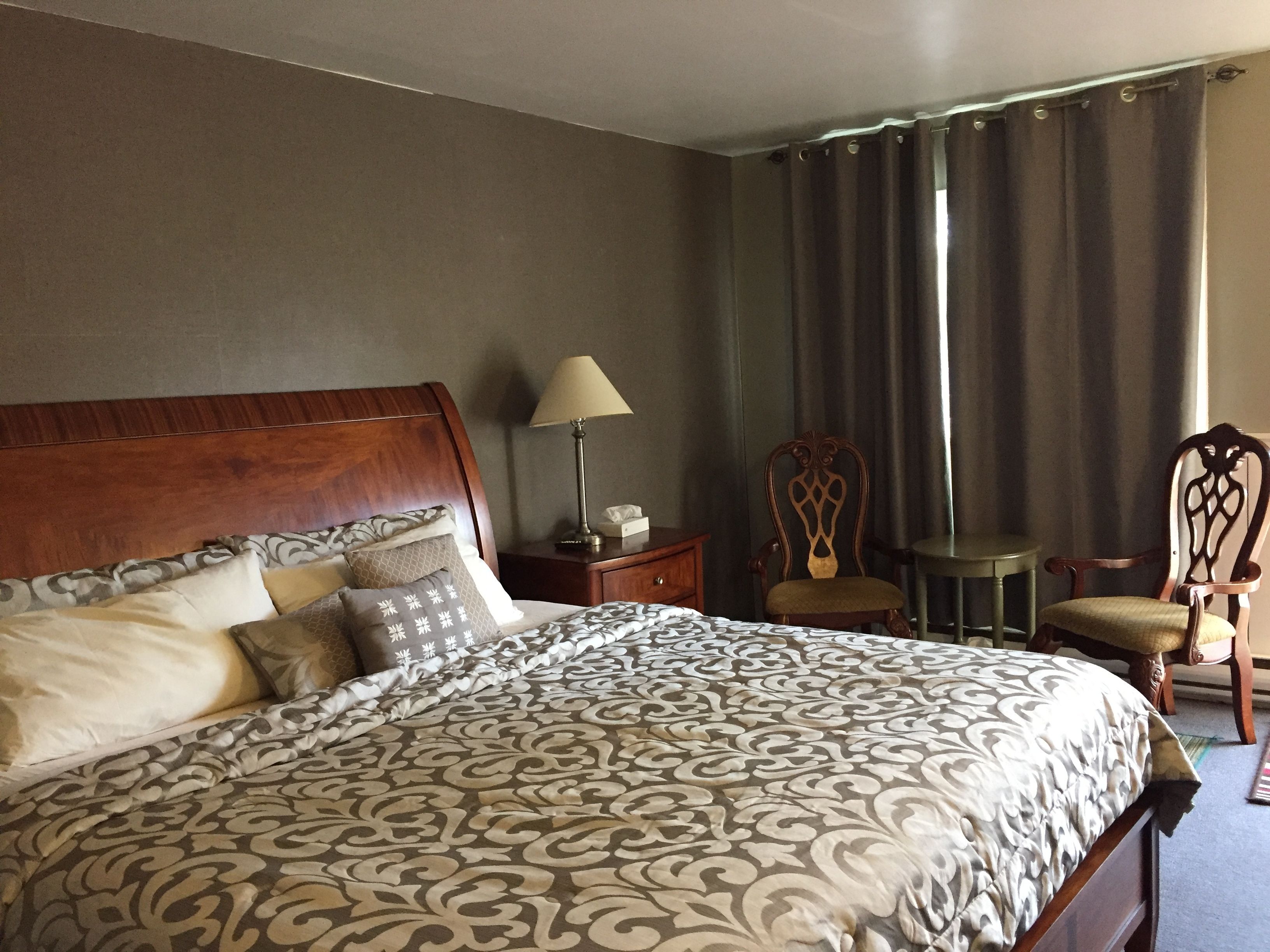 A neatly made bed with a patterned bedspread, a wooden headboard, and a bedside table with a lamp and tissues. There are two chairs and a small table in the room.