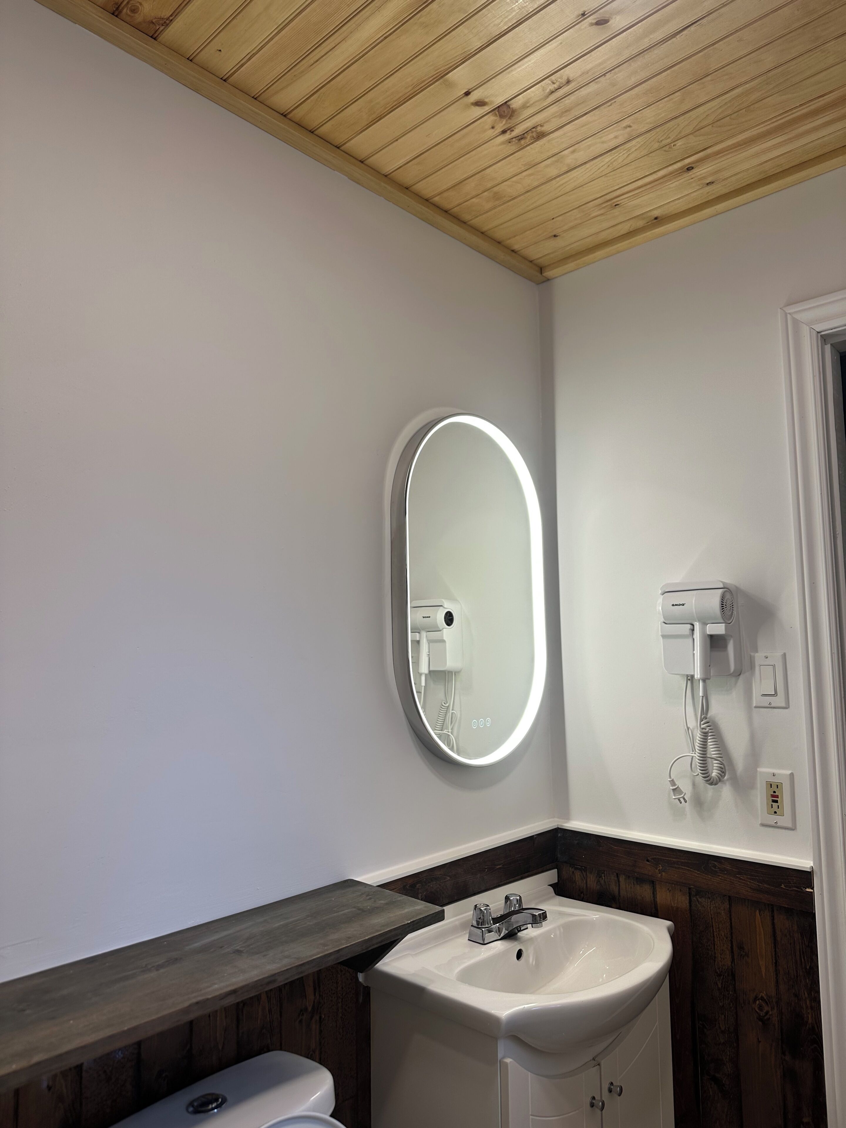 Une salle de bain avec un plafond en bois, un lavabo blanc, un miroir avec un éclairage intégré et un sèche-cheveux.