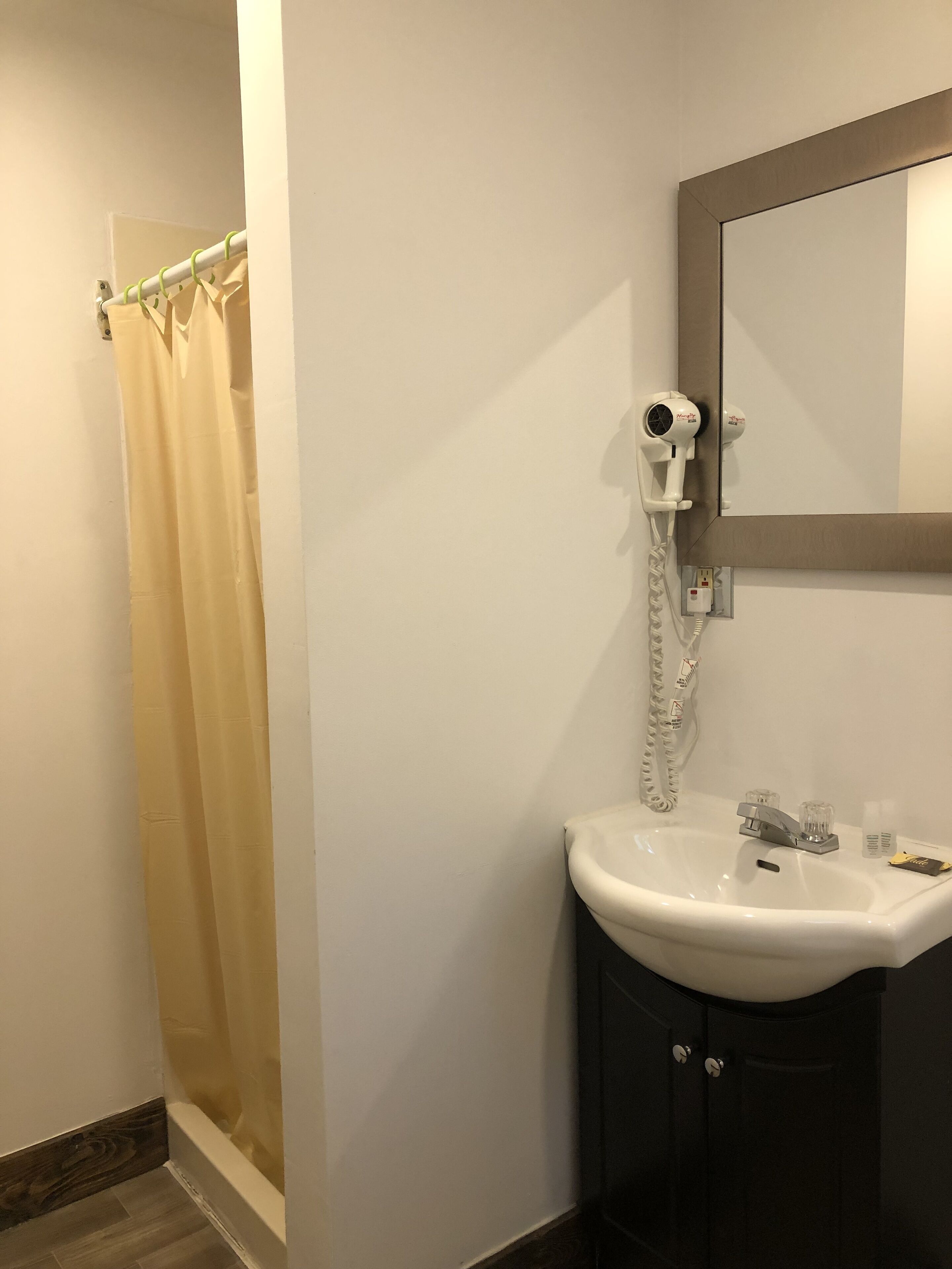 A bathroom with a beige shower curtain, a white hairdryer on a wall-mounted stand, a white sink with a mirror above it, and a dark-colored cabinet below.