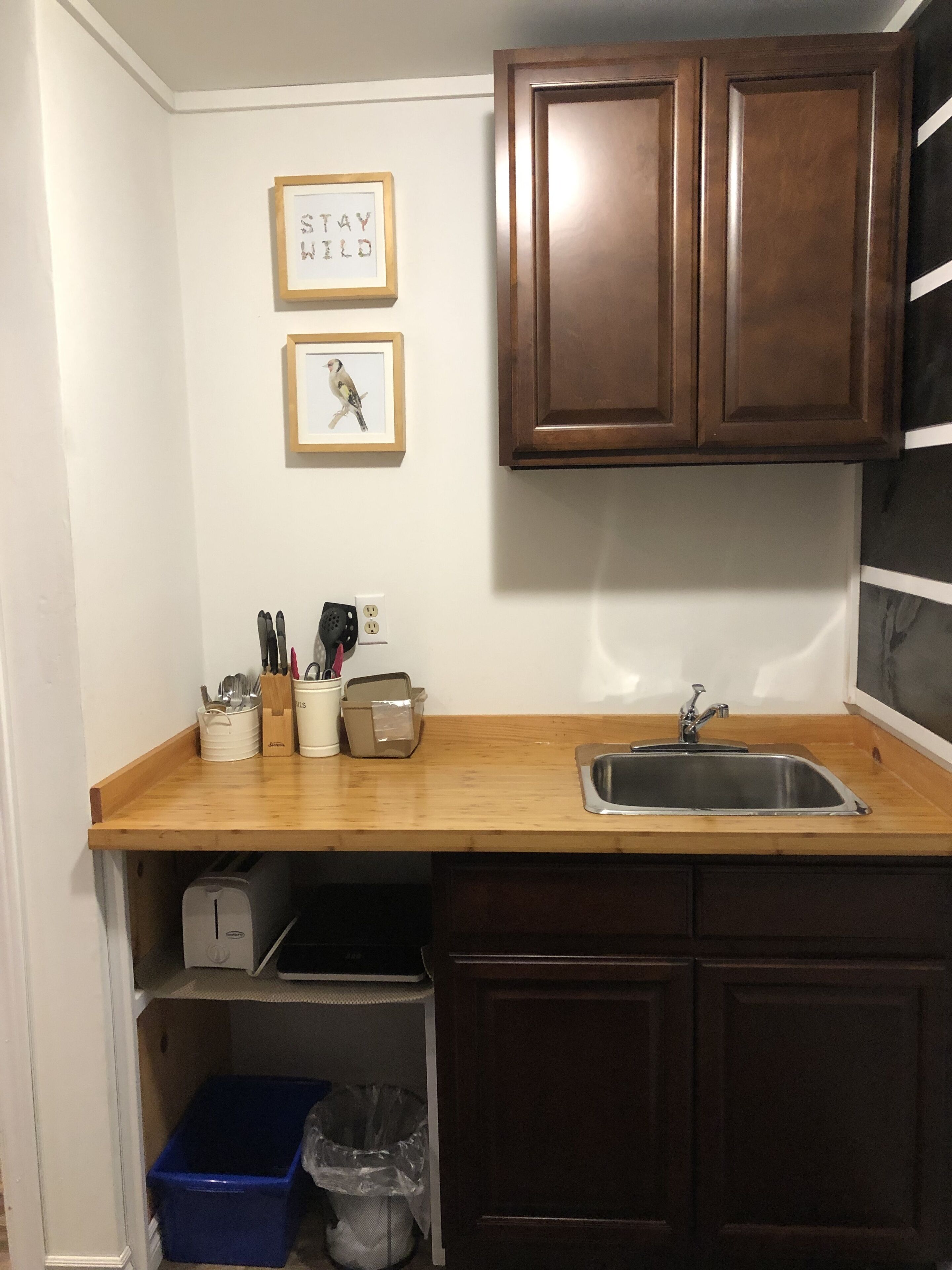 Una cocina compacta con armarios de madera, un lavamanos de acero inoxidable y una encimera con utensilios de cocina. Hay dos cuadros en la pared, uno con texto y otro de un pájaro.