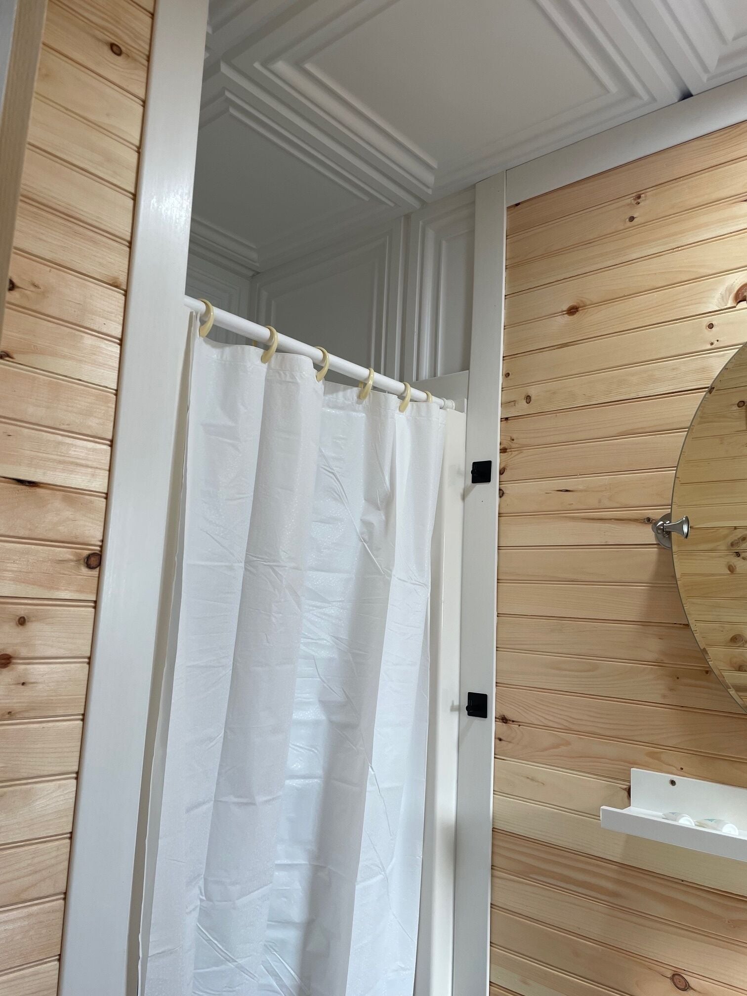 A bathroom with wooden paneling, a white shower curtain, and a round mirror.