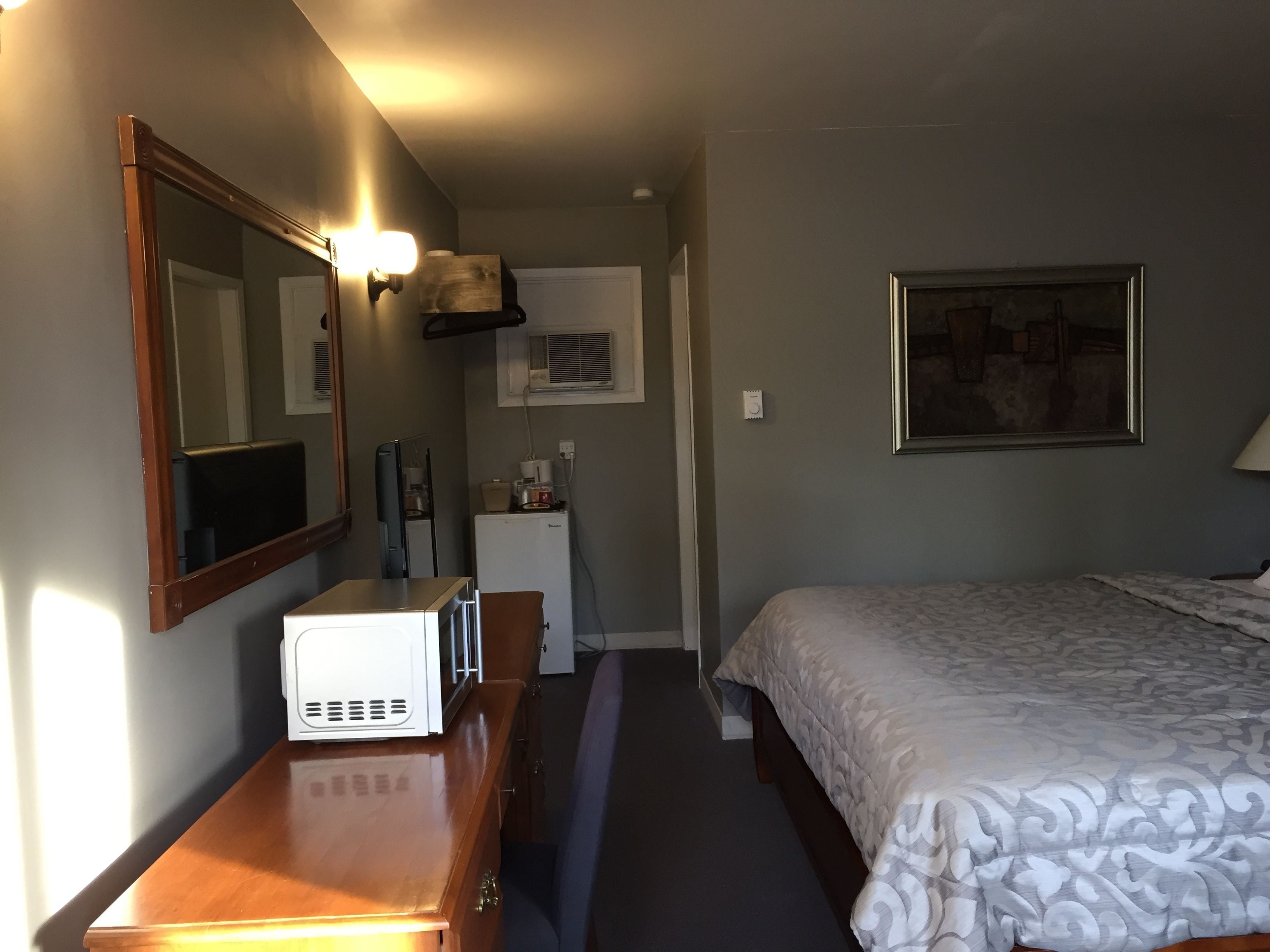 A compact hotel room with a bed, a desk with a microwave, a dresser, a mirror, a framed picture on the wall, and a small kitchenette with a refrigerator and a coffee maker.