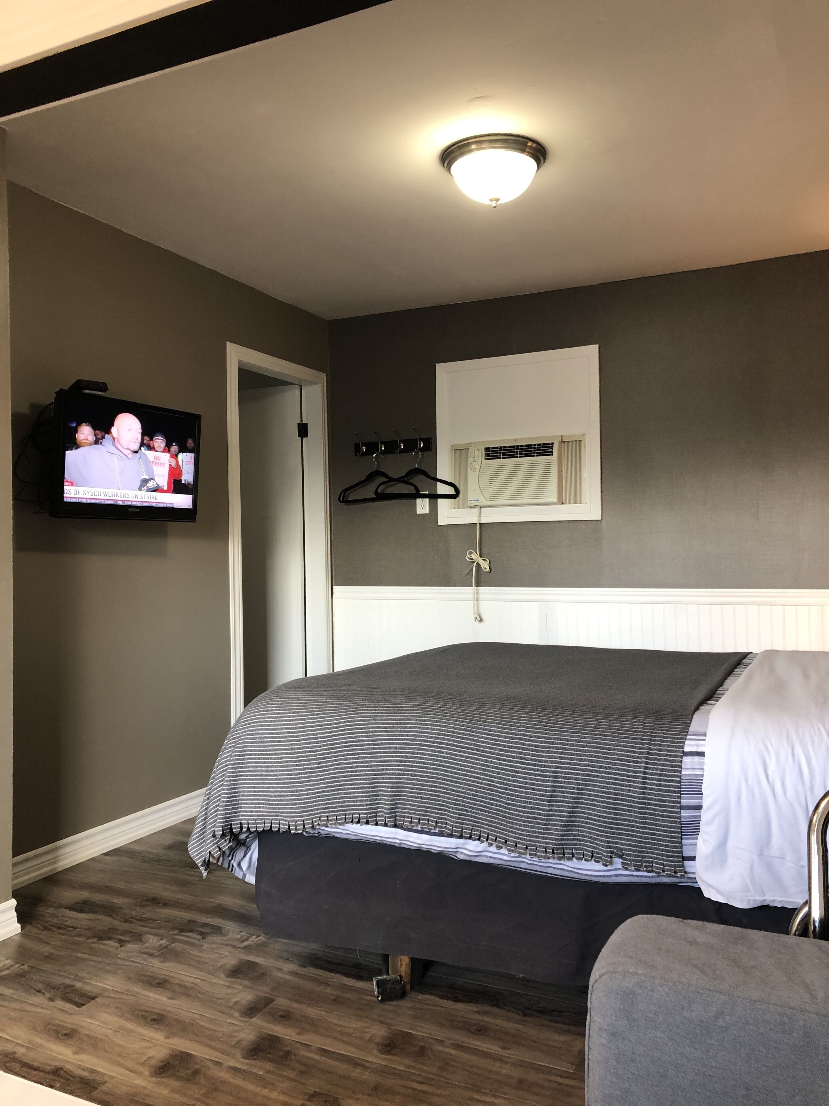 A bedroom with a bed, a television mounted on the wall, a window with blinds, a coat rack, and a small air conditioning unit.