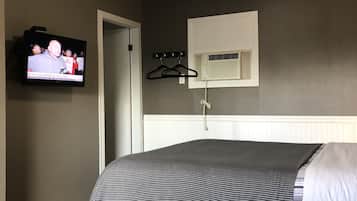 A bedroom with a bed, a television mounted on the wall, a window with blinds, a coat rack, and a small air conditioning unit.