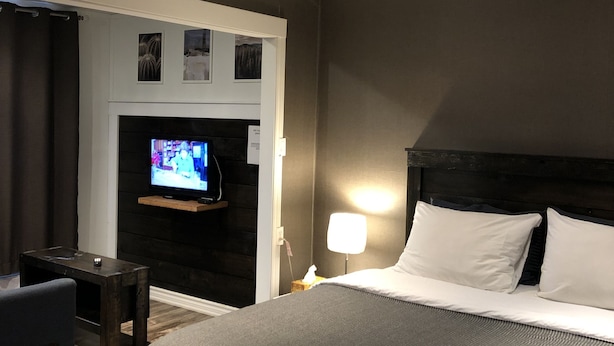 Habitación de hotel con una cama grande, un televisor en una pared oscura, una mesita pequeña y una silla.