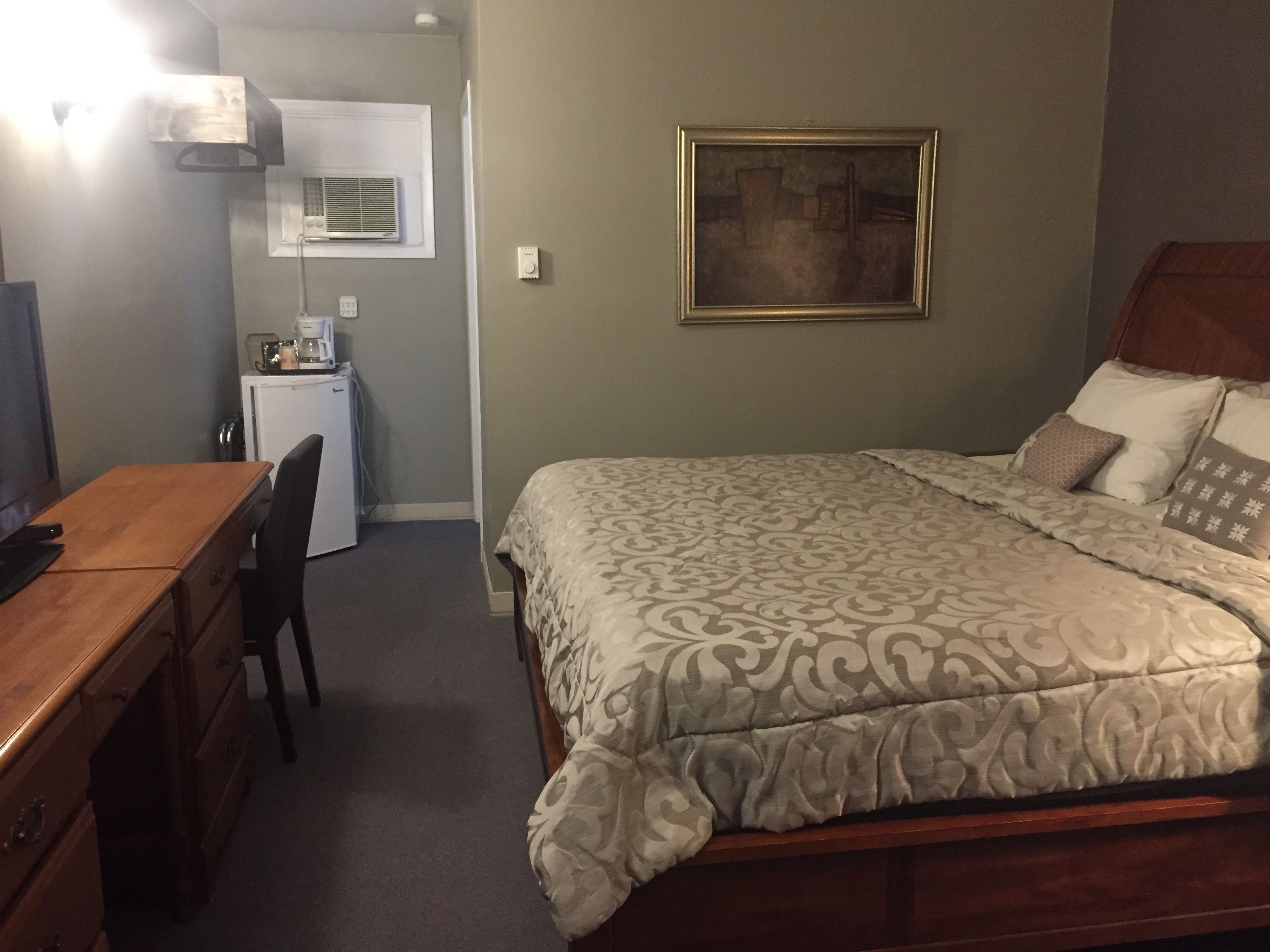 A hotel room with a bed, a wooden desk, a chair, a small kitchenette, and a framed picture on the wall.