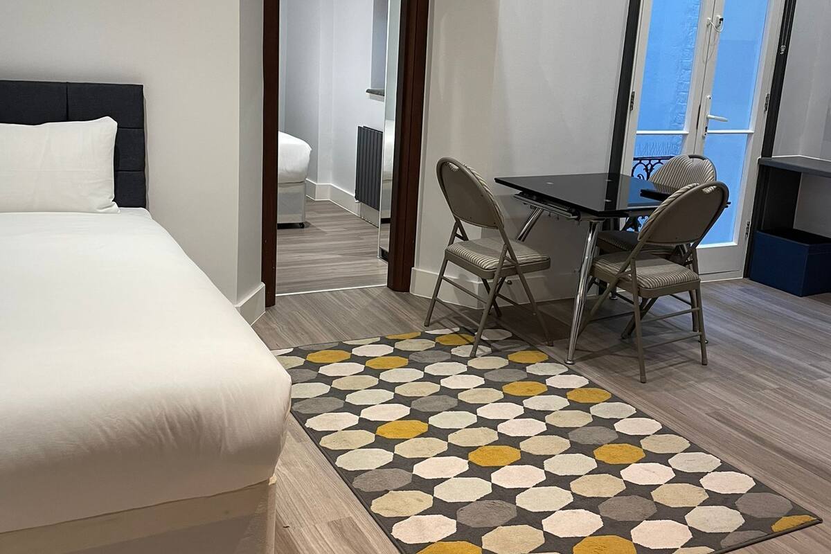 A modern hotel room with a large bed, a dining table with two chairs, a patterned rug, and a view of the outside through a door.