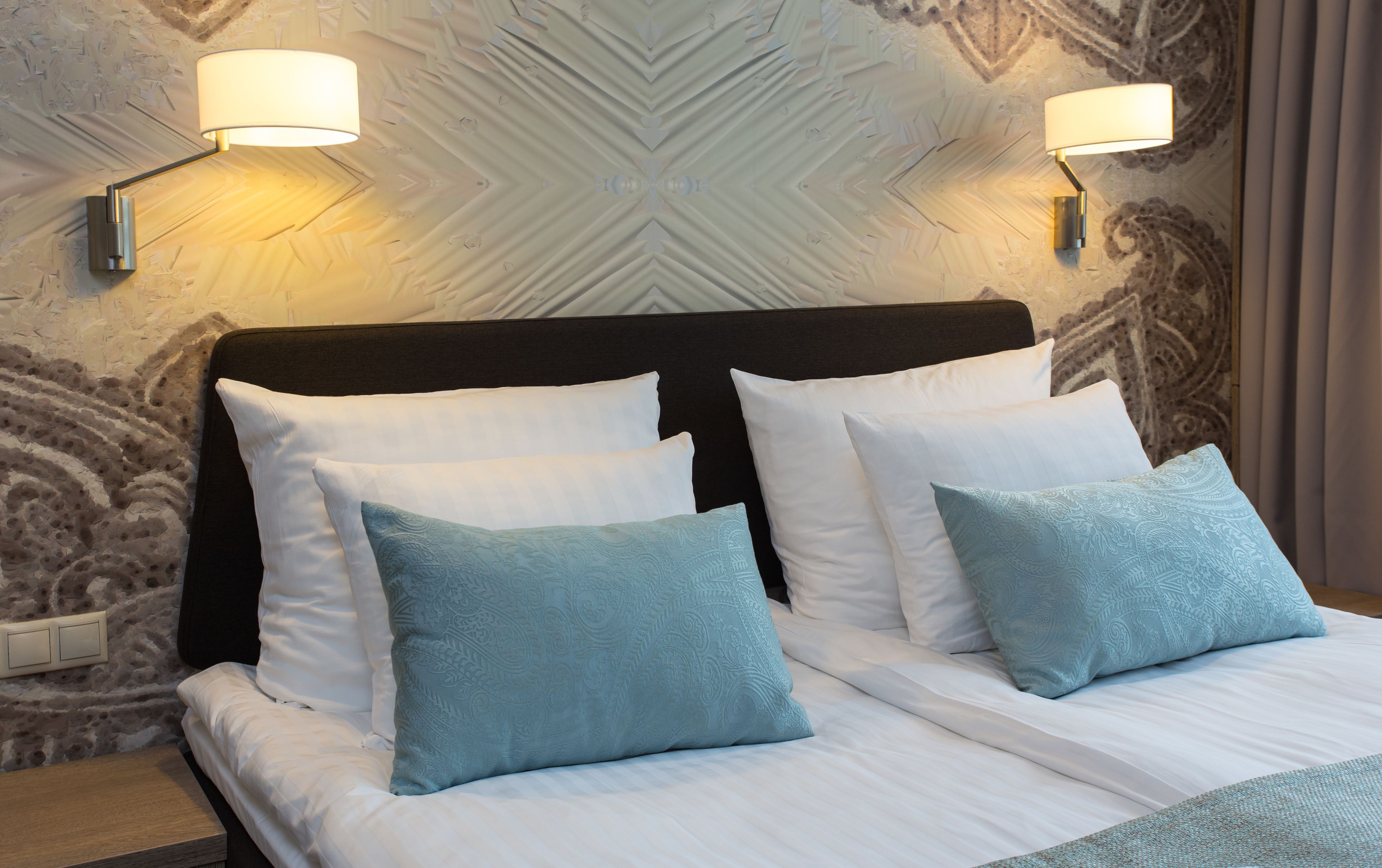Una cama bien tendida con almohadas blancas y azul claro, cabecera oscura y dos lámparas fijadas a la pared.