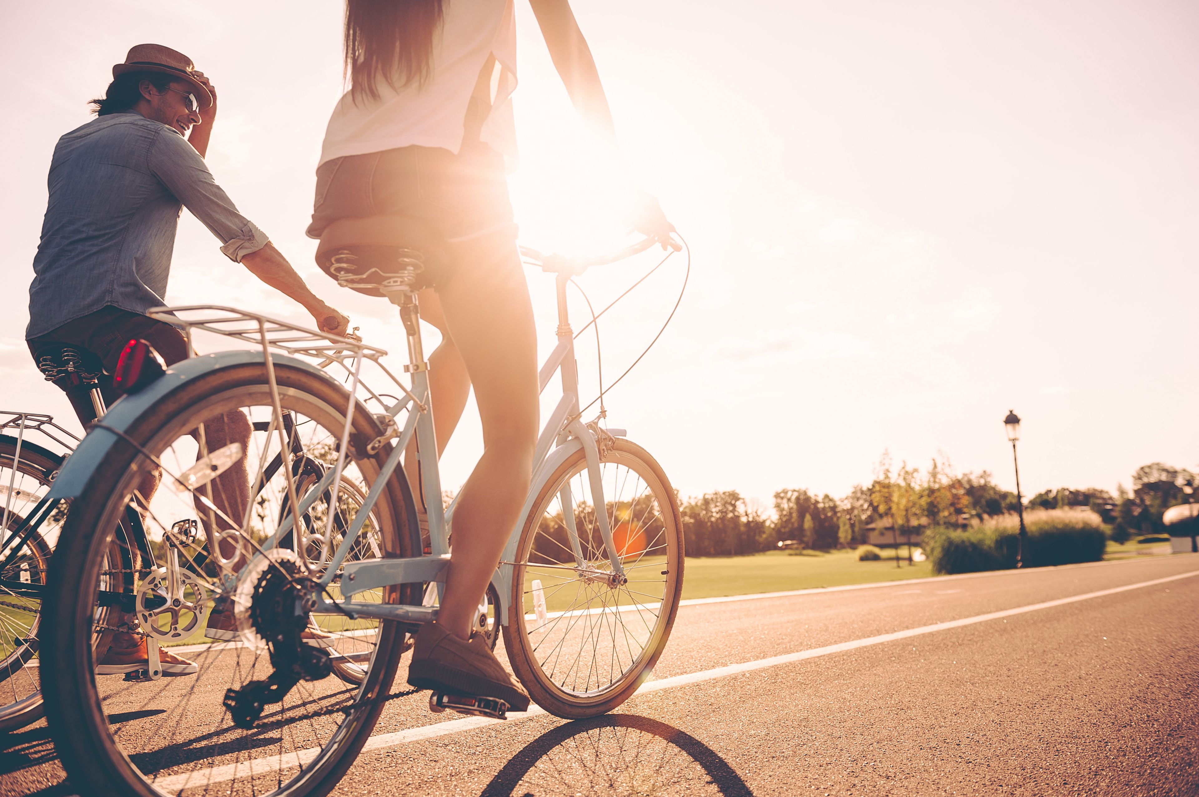 Duas pessoas andando de bicicleta numa estrada pavimentada, com um lampião e vegetação ao fundo.
