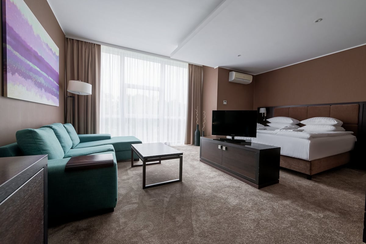 A hotel room with a bed, a green sofa, a TV, a desk, and a large window with curtains.