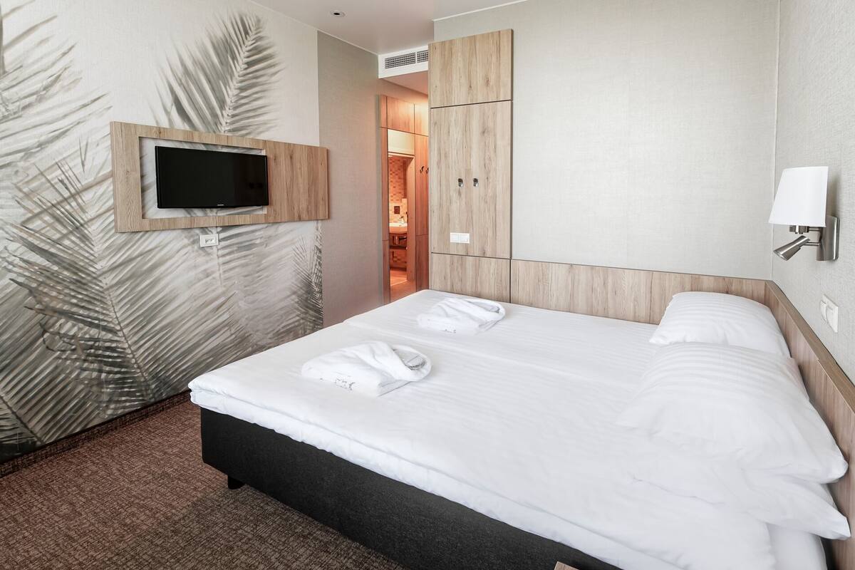 A hotel room with a bed, a TV mounted on the wall, a wooden wardrobe, and a bathroom visible through an open door.