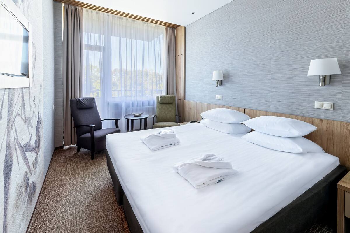 A hotel room with a large bed, two bedside tables, a chair, a small table, and a window with curtains.