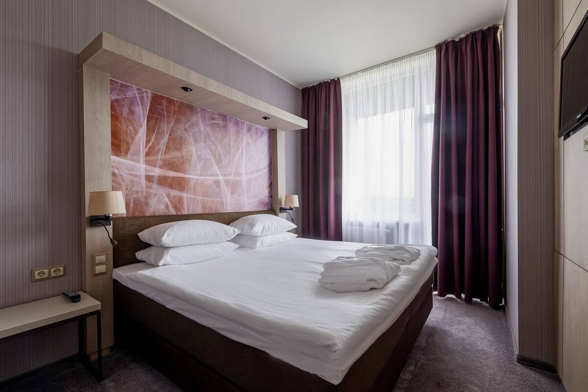 A hotel room with a large bed, two bedside lamps, a TV, and a view of the outdoors through sheer curtains.